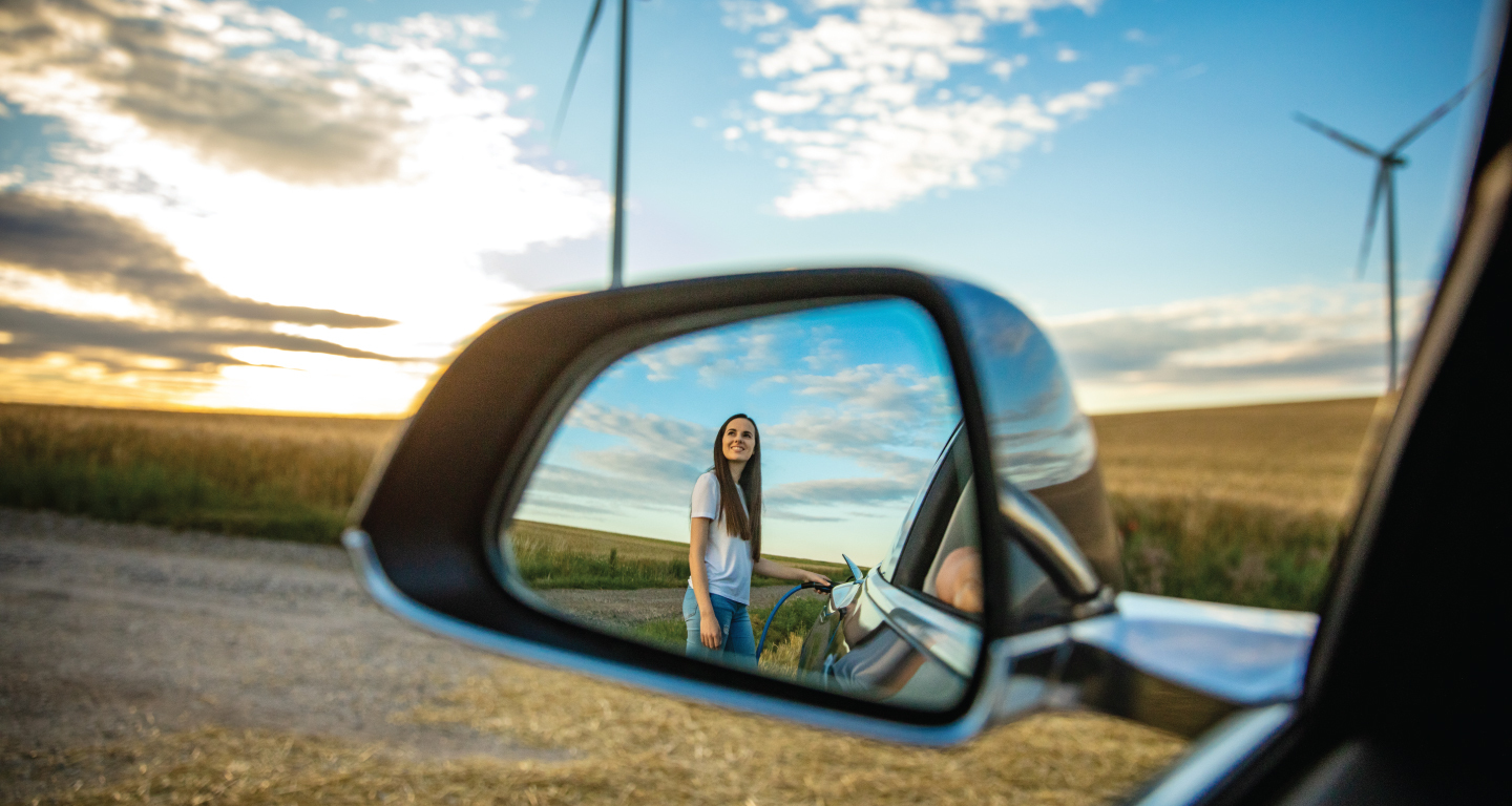 Una donna in piedi riflessa nello specchietto retrovisore di un'auto elettrica in ricarica. Sullo sfondo sono presenti delle pale eoliche 